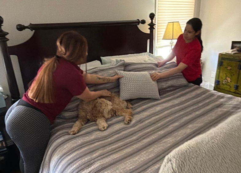 Two women making a bed with a cat lying on it.