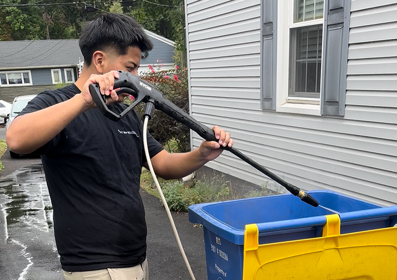 A person cleaning a blue recycling bin with a hose outside a house.