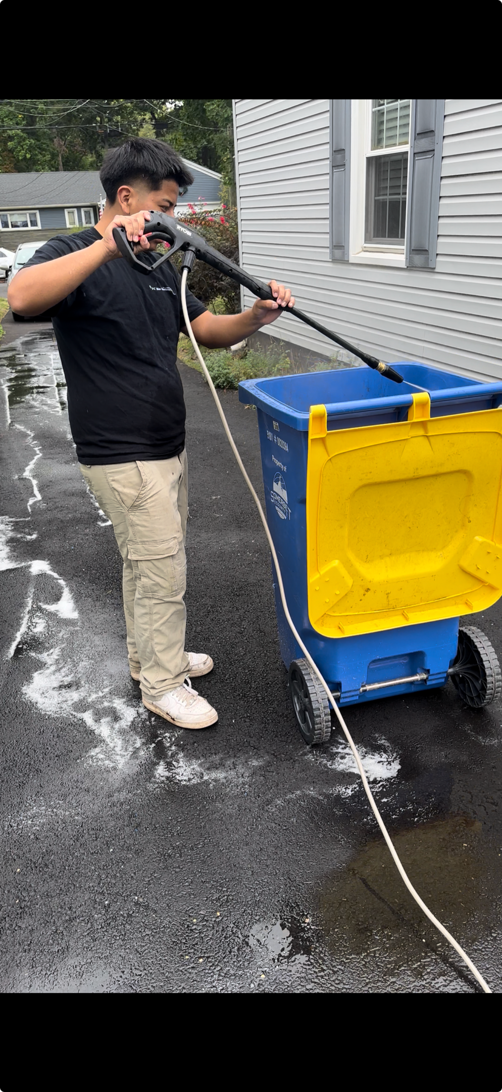 Person cleaning outdoor pavement using a foam sprayer and blue trash bin.